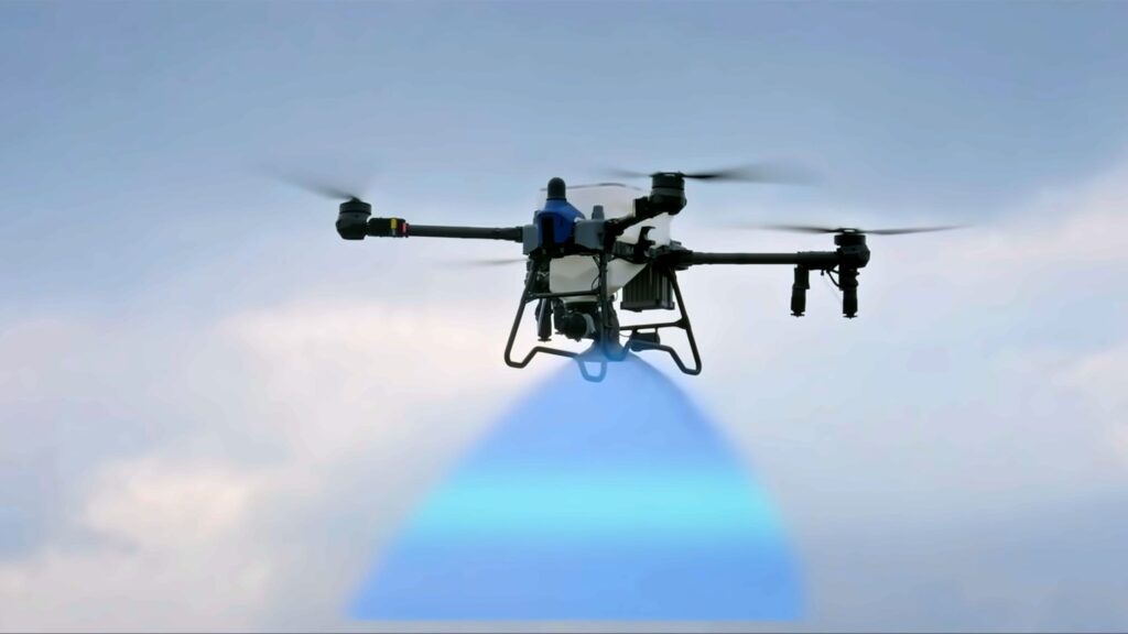 Agricultural drone spraying a field with a downward-facing nozzle and visible spray.